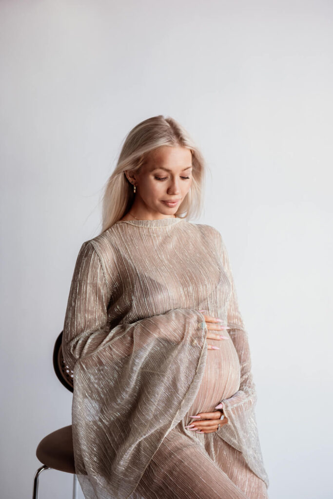 pregnant woman sitting on chair white seamless background and hands on belly facing towards the light photographed by Austin maternity photographer kat harris