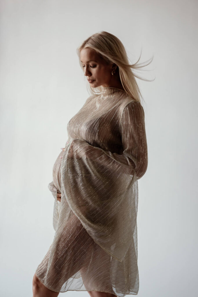 Pregnant woman in flowy gold sheer dress with hands on belly and windblown hair photographed by Austin maternity photographer Kat Harris