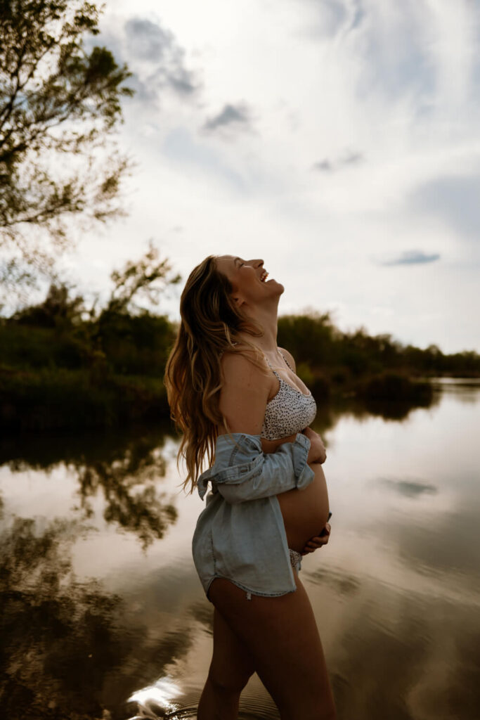 Moody pregnancy portrait of pregnant mother at the river in her bathing suit with a denim button-up blouse falling off her shoulders while laughing photographed by Austin maternity photographer Kat Harris.