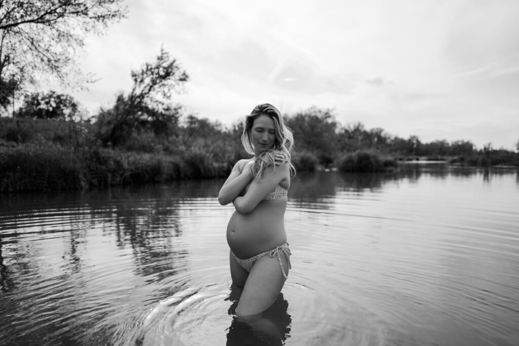 chic black and white maternity portrait of woman in a bathing suit in the water holding herself with her hair over one shoulder photographed by Austin fine art maternity photographer Kat Harris.