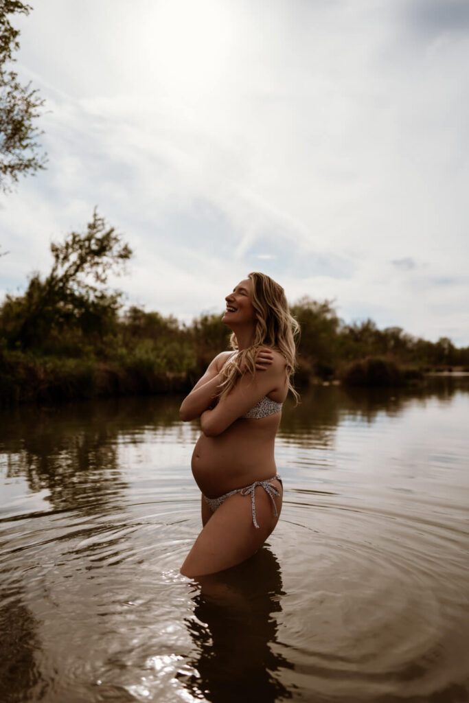 Moody pregnancy portrait of woman in a bikini in the water hugging herself with her eyes closed smiling towards the sun photographed by Austin portrait photographer Kat Harris.