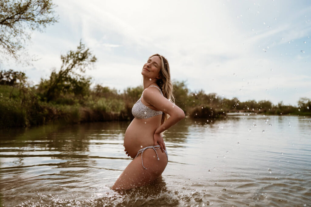 Profile view of pregnant mom with one hand on her belly and the other on her lower back standing in a peaceful river with water splashing by her photographed by Austin maternity photographer Kat Harris.