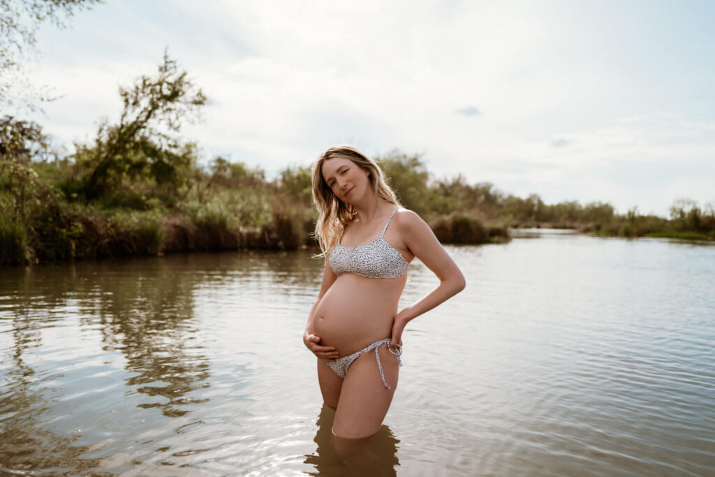 Pregnant woman softly holding her baby bump in the water at golden hour in her bathing suit photographed by Austin maternity photographer Kat Harris.