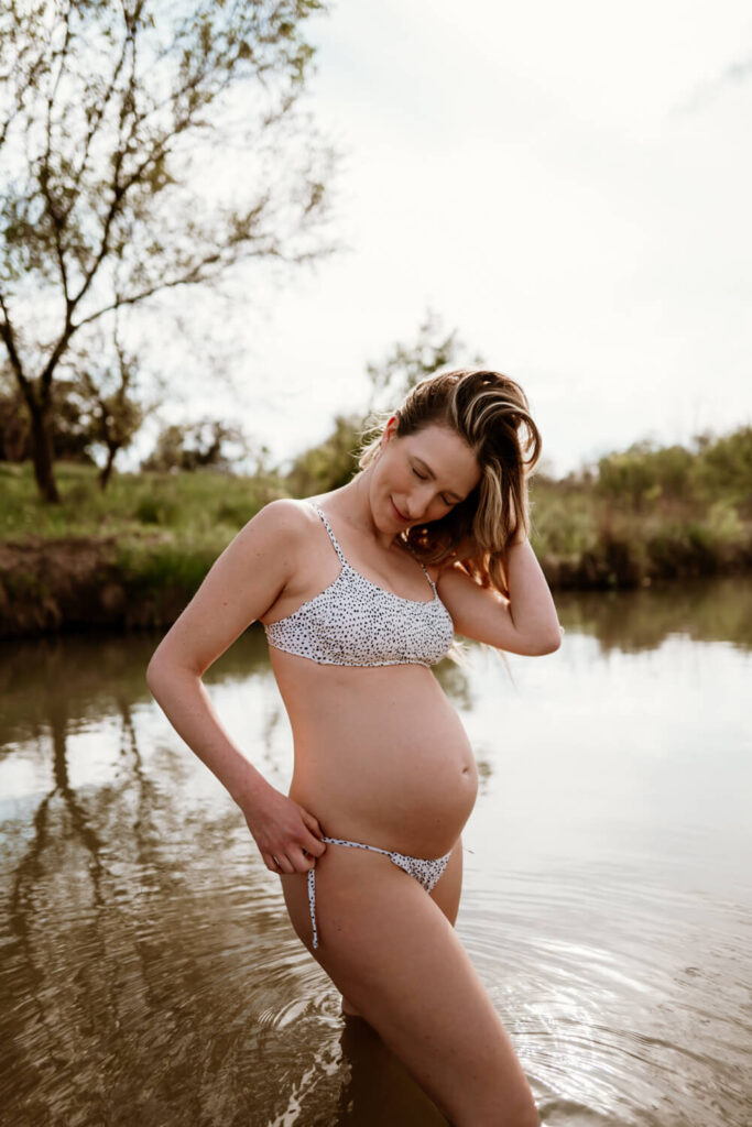 Lake maternity portraits of pregnant woman in her bathing suit fixing her hair in the water photographed by Austin pregnancy photographer Kat Harris.