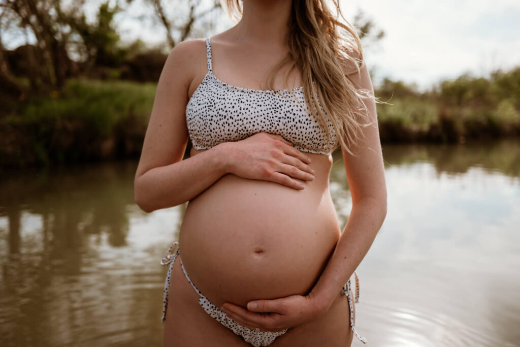 close up photo of a pregnant woman in a bikini lovingly holding her belly while she wades in Town Lake photographed by Austin water maternity photographer Kat Harris.
