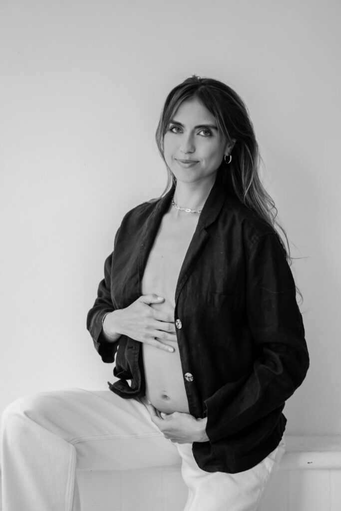 Classic black and white portrait of pregnant mother with hand on her baby bump seated in white light studio photographed by Downtown Austin maternity photographer Kat Harris.