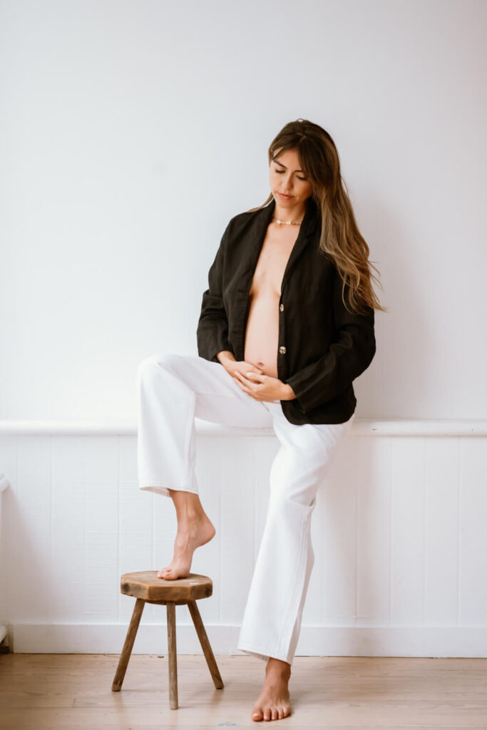 Pregnant woman holding her baby bump looking downward with her foot on a stool at white light studio in Downtown Austin, Amoroso Studio, photographed by Austin portrait and maternity photographer Kat Harris.