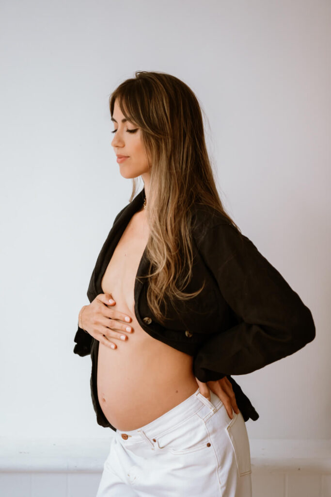 side profile portrait of pregnant woman with her hand on her belly and her hair swept over her shoulder at in front of a white wall at downtown Austin natural light Amoroso Studio photographed by Austin maternity portrait photographer Kat Harris.