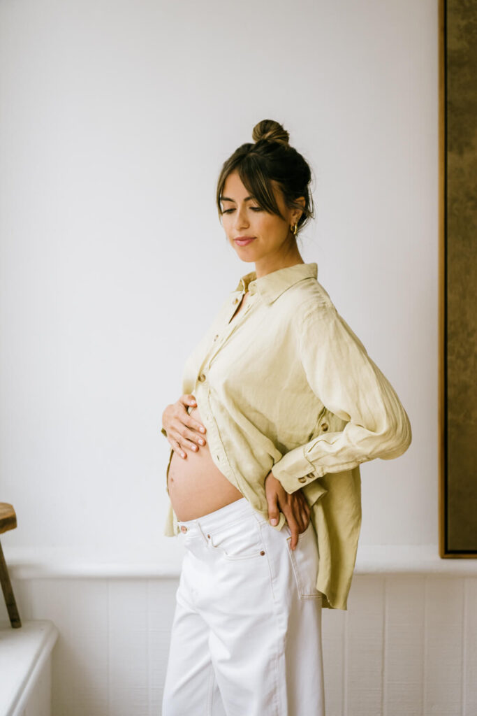 Chic pregnant mother with high messy bun an unbuttoned tan blouse and baggy white jeans and hand on her belly at natural light Downtown Austin studio photographed by Austin maternity photographer Kat Harris