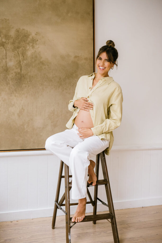 Photo of laughing pregnant mother sitting on a stool with tan button-up at natural light studio in Downtown Austin by studio portrait photographer Kat Harris.