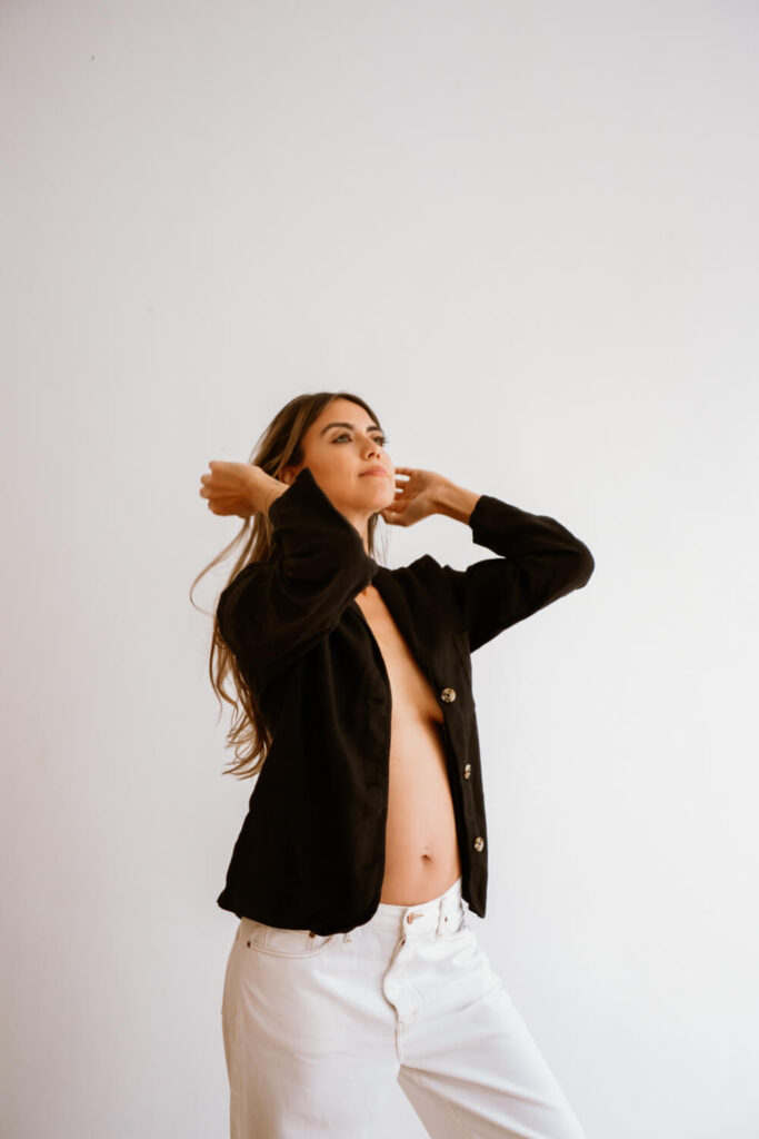 Pregnant mamma fixing her hair with black blazer and baby bump in front of a white wall at Downtown Austin studio by natural light Austin maternity photographer Kat Harris.