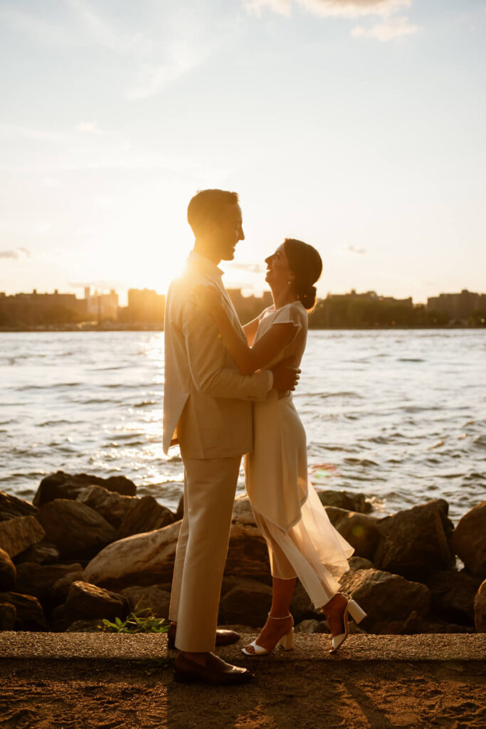 Brooklyn engaged couple embraces at the waters edge in Williamsburg Brooklyn laughing and smiling at the peak of golden hour with the Manhattan skyline behind them.