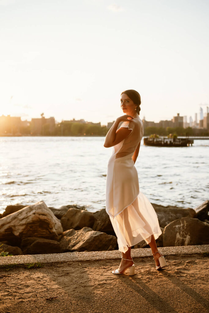 Chic bride in custom wedding dress stands at Brooklyn's water's edge at sunset and looks over her shoulder with the stunning Manhattan skyline lit up in golden hour.