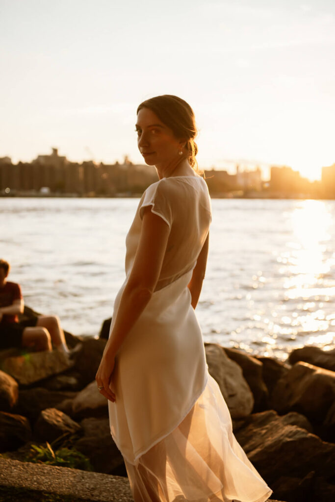 Portrait of a bride with her red hair up in a bun looking over her shoulder at the camera with the golden sunset and Manhattan skyline in the background.