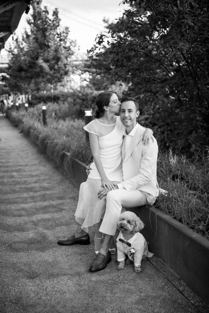 engagement photo shoot with a dog at Domino Park in Williamsburg Brooklyn photographed by Austin wedding photographer Kat Harris with bride sitting on fiancé's lap kissing his forehead with the puppy smiling towards the camera.