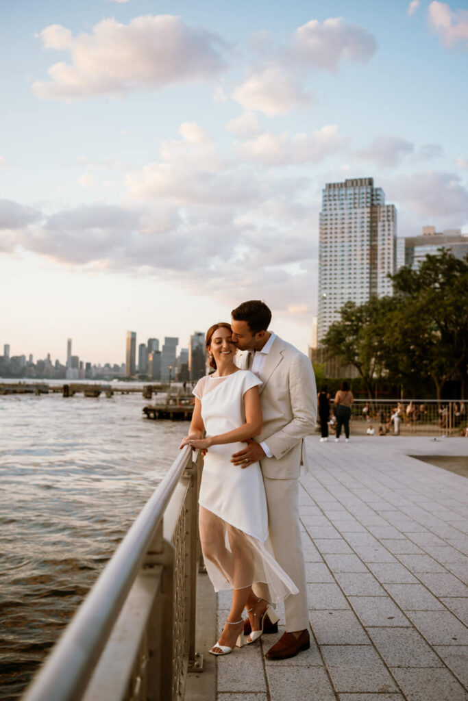 Woman in custom chic white dress stands and smiles at the waters edge of Domino Park at sunset and receives a kiss from her dapper fiancé. 