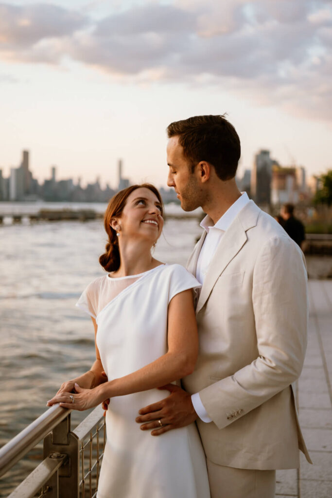 Strolling engaged couple stop at the waters edge of Brooklyn's Domino Park and bride looks happily at her handsome fiancé during golden hour.