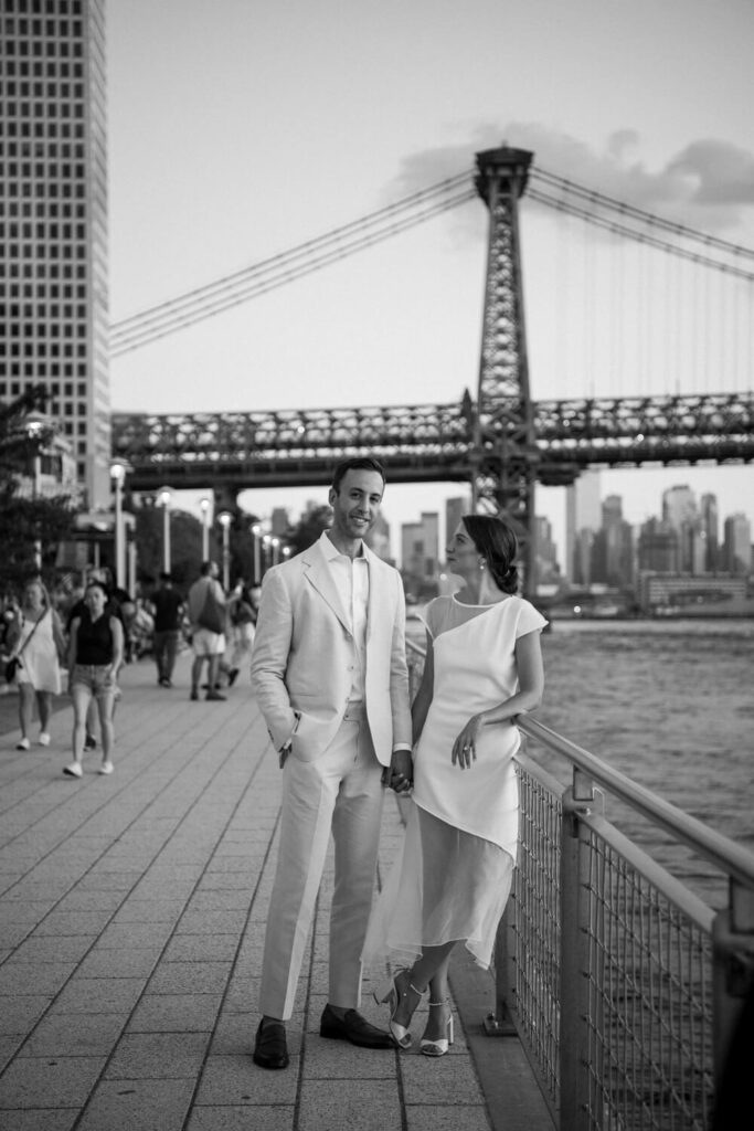 Chic Brooklyn couple poses for an engagement portrait at Domino Park with the bride in a white chic dress looking lovingly towards her fiancé while he smiles towards the camera.