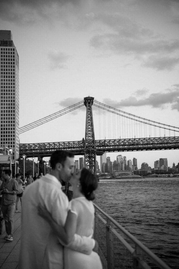 engaged couple pause for a kiss on the Domino Park walkway at sunset with the Williamsburg bridge and the Manhattan skyline in the background.