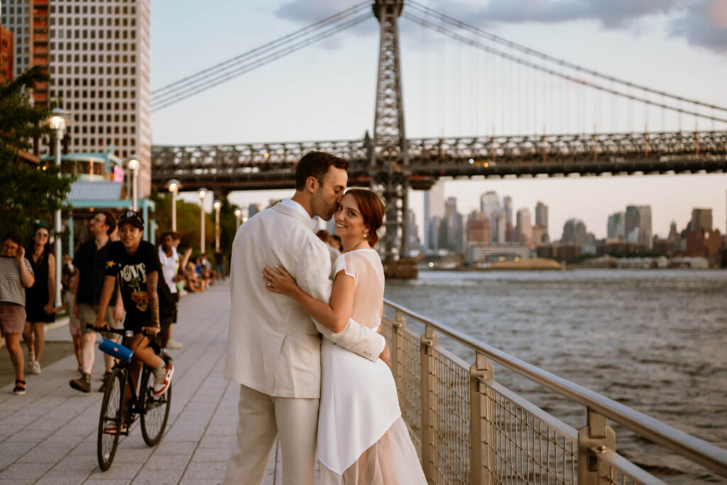 In the midst of the bustle of a summer evening at Wiliamsburg's Domino Park, Brooklyn engaged couple take a romantic stroll at sunset and pause for a kiss while surrounded by locals and bikers with Williamsburg Bridge in the background.