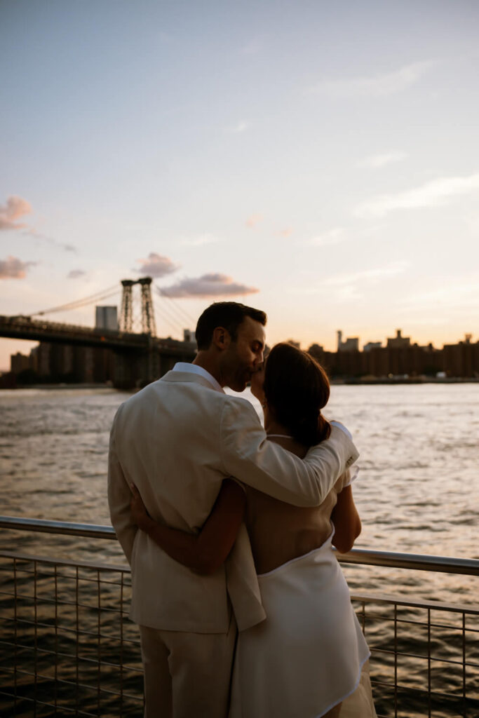 As sunset turns to dusk at Domino park engaged Brooklyn couple looks to the Manhattan skyline and steal a quiet moment together and a kiss during their engagement photos.