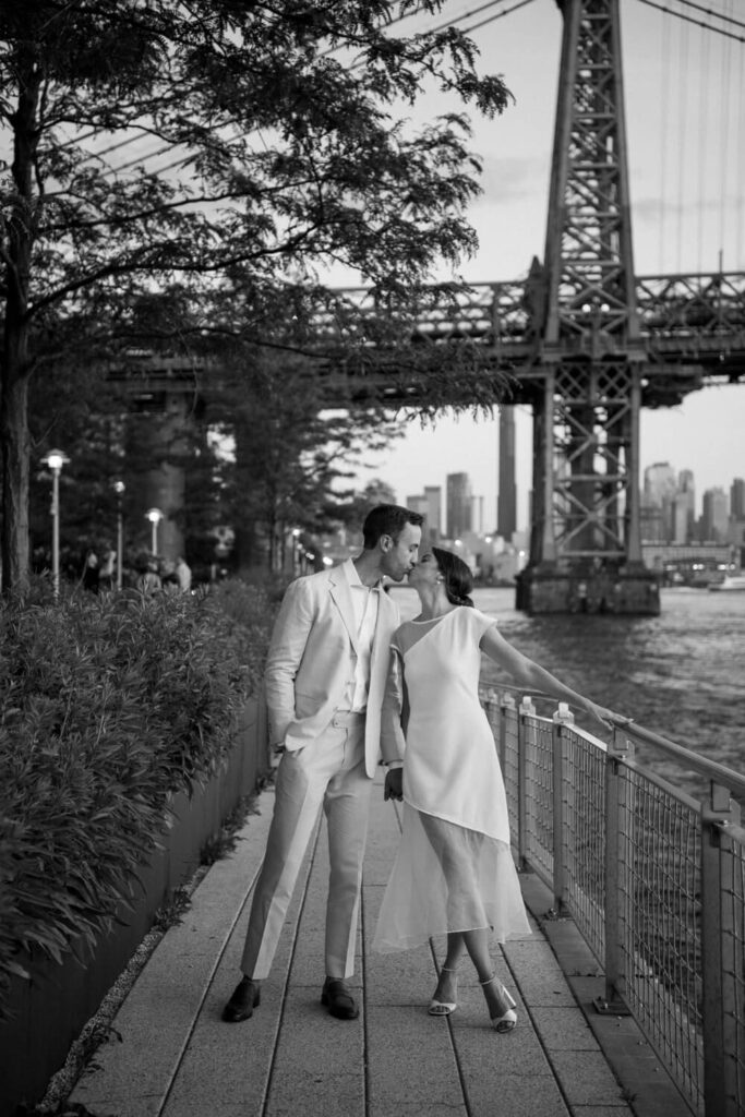 Bride and groom take a leisurely walk at Domino Park at dusk during engagement photos and pause for a quick kiss with the Williamsburg Bridge in the background.