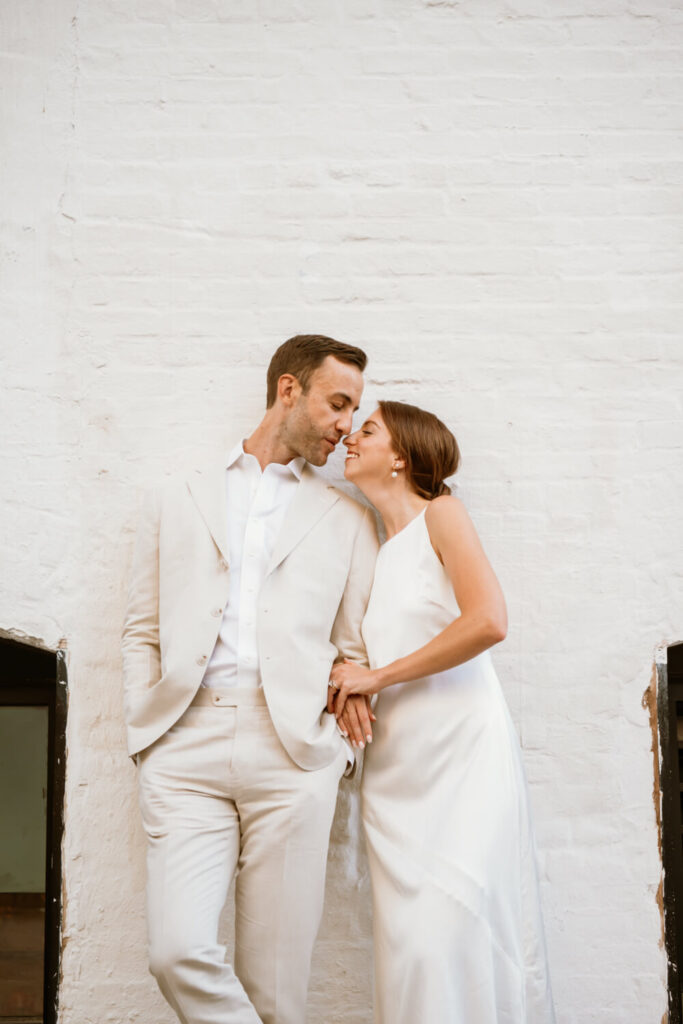 Brooklyn couple lovingly leaning in for a kiss during their engagement portraits in Williamsburg against a white brick wall backdrop.