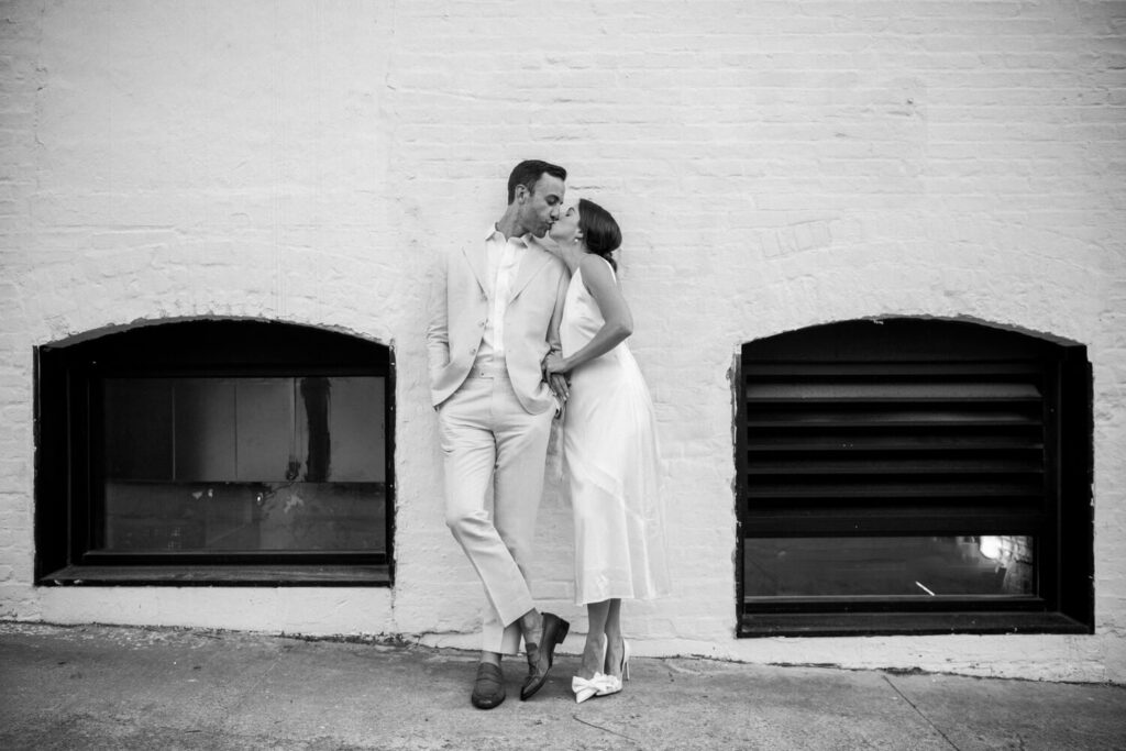 woman in white dress kissing her fiancé in a tan suit against a white brick wall in timeless engagement portraits in Williamsburg Brooklyn.