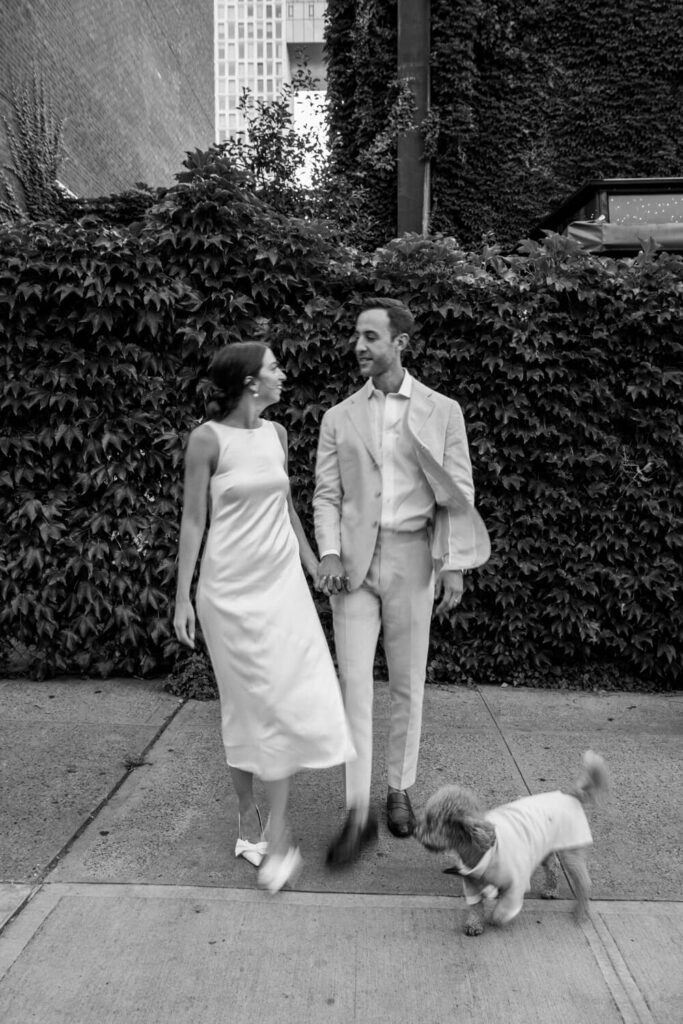 Woman in white dress and heels going on a walk with her fiancé and their dog smiling towards each other in front of Aurora restaurant during their Brooklyn engagement session.