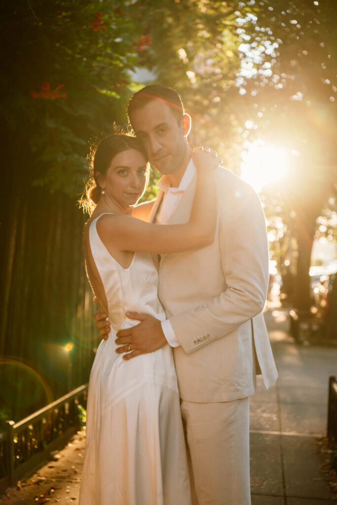 Brooklyn engaged couple embracing on a Williamsburg sidewalk with the golden sunset light behind them coming through the green trees.