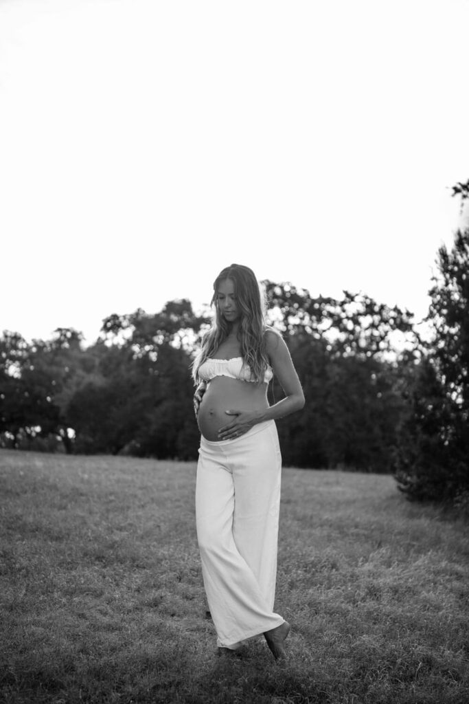 Austin maternity photo of pregnant woman in a white two piece set holding her baby bump looking downward in a wide open field photographed by Austin maternity photographer Kat Harris.