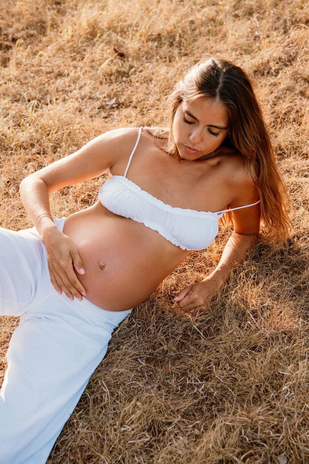 Austin maternity mamma dressed in white linen bralette and white pants laying in a field holding her belly photographed by Austin maternity photographer Kat Harris.