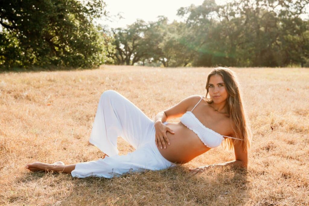 natural light portrait during golden hour in a field of a pregnant woman holding her baby bump while lying down on the ground photographed by Austin maternity photographer Kat Harris.