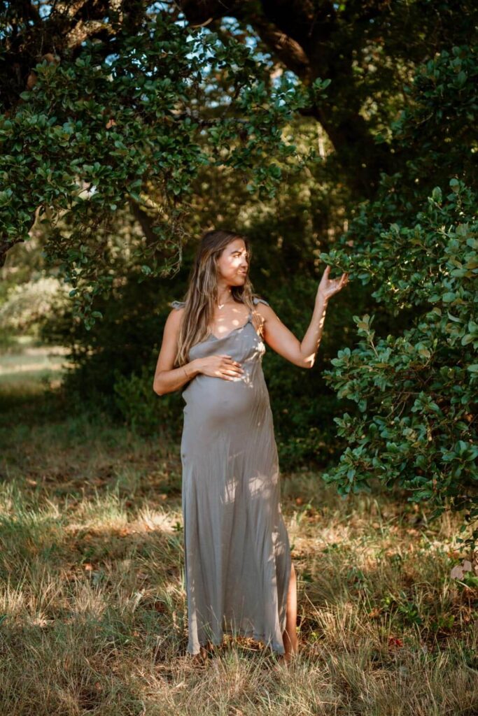 Maternity photoshoot in nature of pregnant woman holding her baby bump in grey slip dress while reaching out to touch a tree branch and being dappled with morning light photographed by Austin maternity photographer Kat Harris.