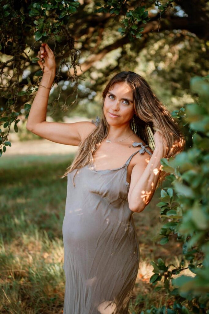 Austin maternity photo of pregnant mom surrounded by trees in a grey slip dress fixing her hair photographed by Austin maternity photographer Kat Harris.