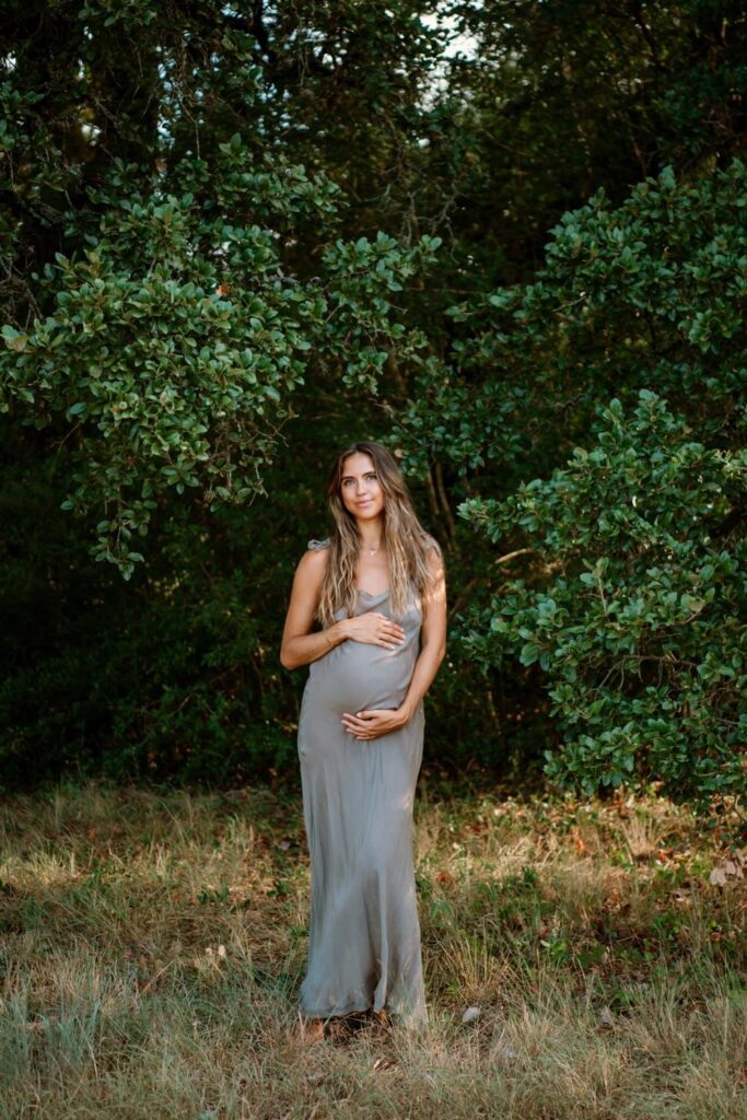 Austin maternity shoot in field with trees with woman in grey slip dress holding her baby bump with long brown hair framing her face photographed by Austin maternity photographer Kat Harris.