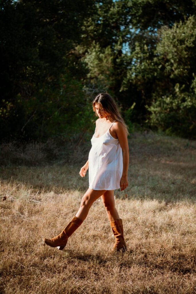 Pregnant woman in white slip dress and brown riding boots walking in a field during golden hour photographed by Austin maternity photographer Kat Harris.