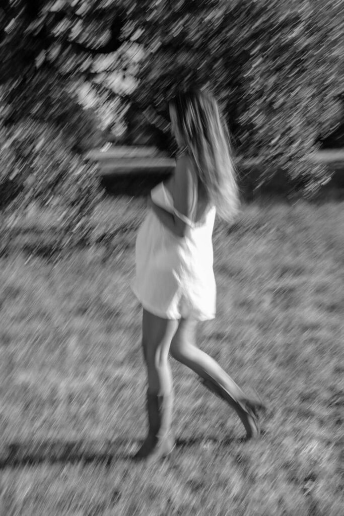 motion blur black and white photo of pregnant woman holding her baby bump walking through a field in a white slip dress photographed by Austin maternity photographer Kat Harris.