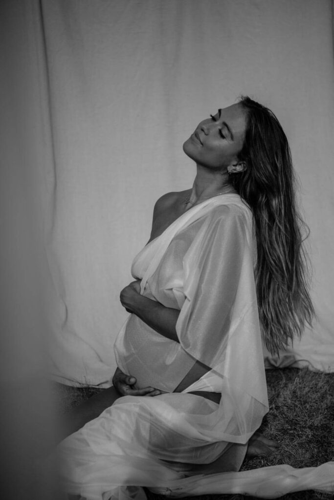 black and white ATX maternity photo of pregnant woman on her knees looking toward the sky with her eyes closed holding her baby bump dressed in sheer white fabric photographed by Austin maternity photographer Kat Harris.