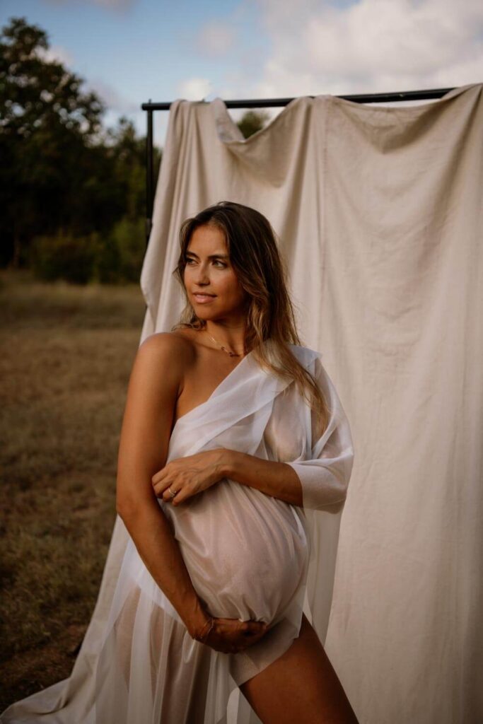 ATX maternity photo with a canvas backdrop in a field and pregnant woman holding her baby bump while draped in white sheer fabric photographed by Austin maternity photographer Kat Harris.