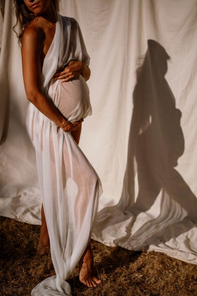 Austin maternity photo with moody golden light of pregnant woman standing in front of a canvas back drop looking downward while holding her baby bump and draped in white sheer fabric photographed by Austin maternity photographer Kat Harris.