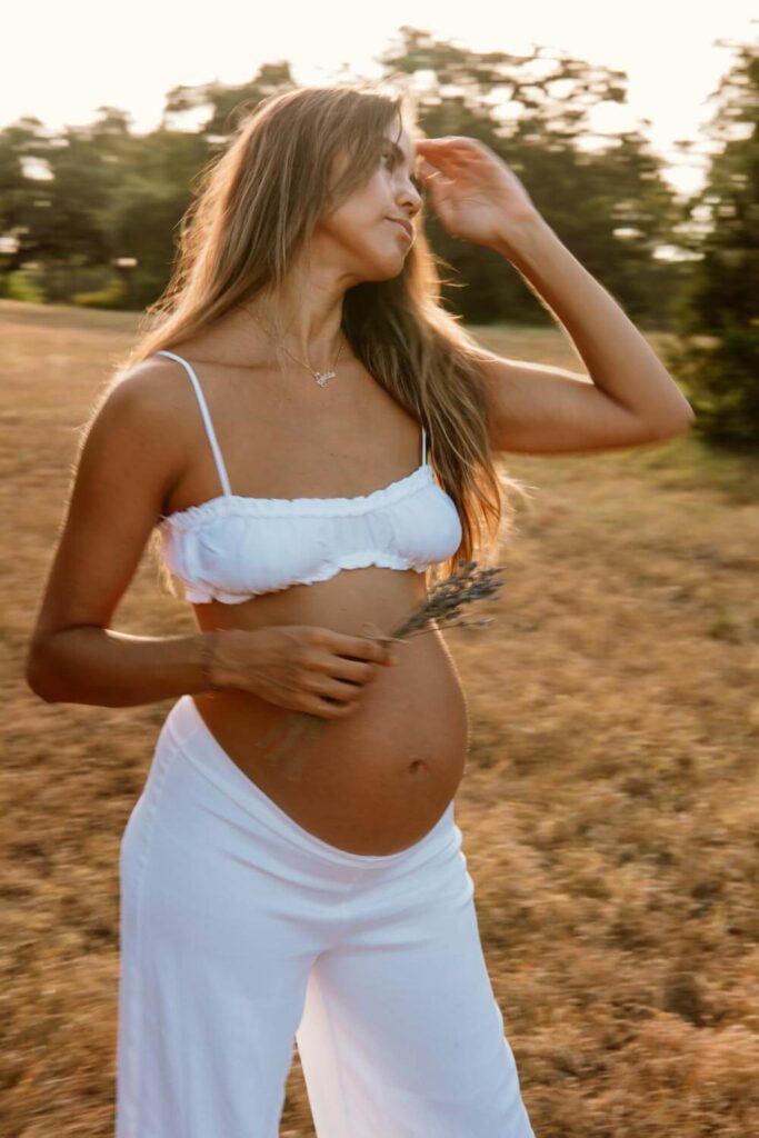 ATX maternity photo of pregnant woman holding fresh lavender while fixing her hair in the morning light in an Austin field in a white two piece set photographed by Austin maternity photographer Kat Harris.