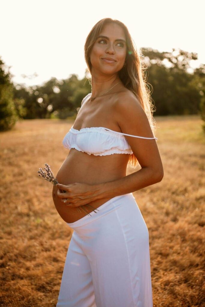 Pregnant woman holding fresh lavender to her bare baby bump dressed in a white linen set in a field photographed by Austin maternity photographer Kat Harris.