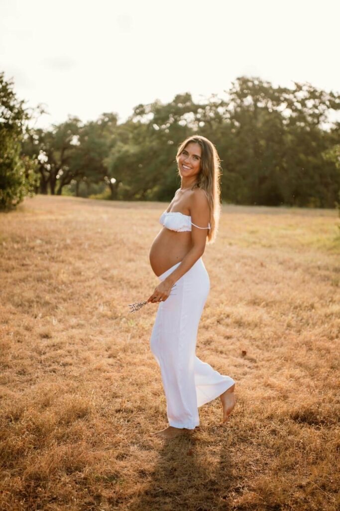 Pregnant woman showing off her baby bump during golden hour in a field smiling back at the camera in a white two piece set photographed by Austin maternity photographer Kat Harris.