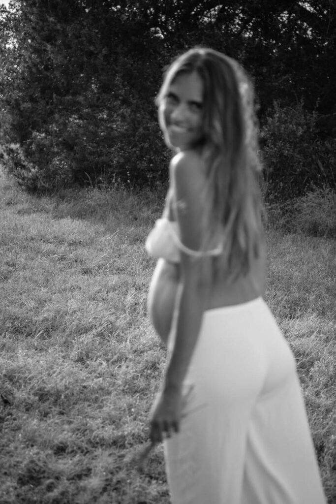 back focused black and white photo of pregnant woman walking away in a field and looking back and smiling towards the camera photographed by Austin maternity photographer Kat Harris.