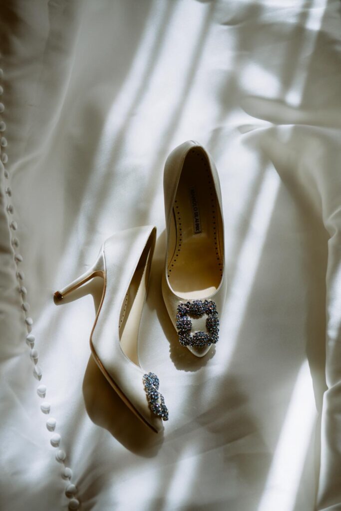 overhead photo of white Manolo Blahnik wedding high heels with blue stones on the tone laid on the train of the wedding gown with moody lighting photographed by New York City wedding photographer Kat Harris.