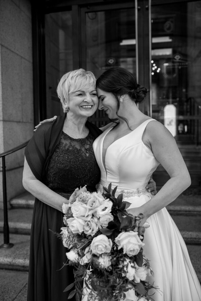 black and white portrait of bride holding her bridal bouquet hugging her mother as she smiles photographed by New York City wedding photographer Kat Harris.