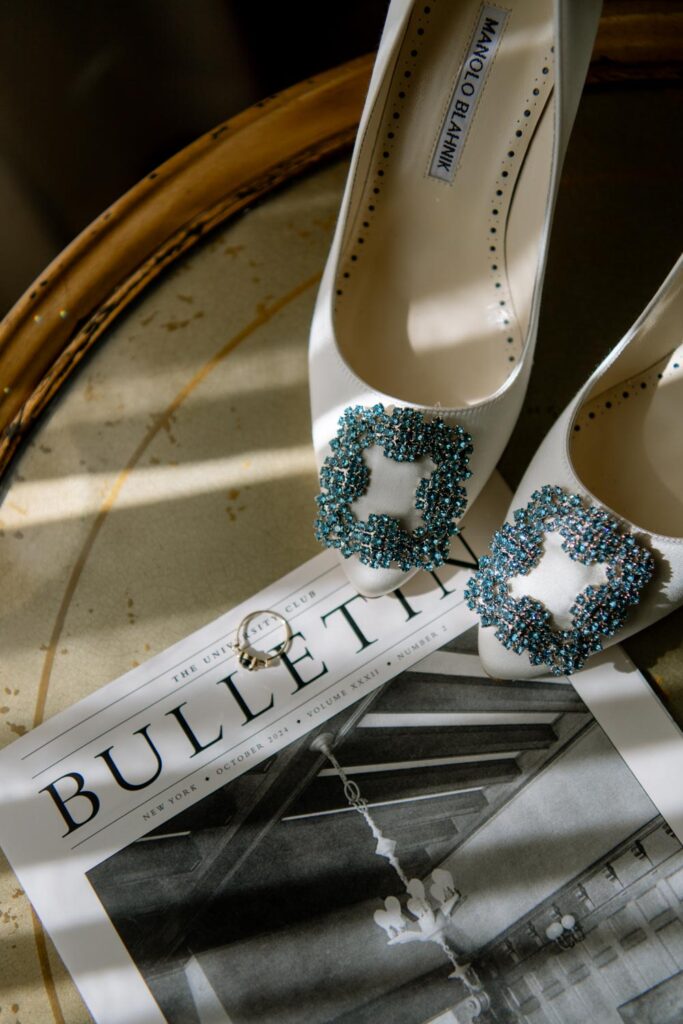 Detail of Manolo Blahnik wedding shoes on a table with the with the brides engagement ring sitting on the daily University Club of NY bulletin with moody lighting photographed by New York City wedding photographer Kat Harris.