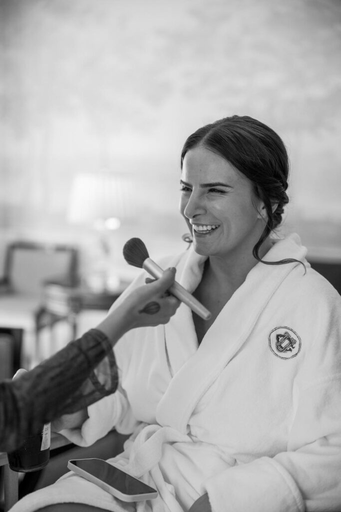 Bride in white robe in the University Club of New York bridal suite laughing as she gets her makeup done in preparation for her wedding photographed by New York City wedding photographer Kat Harris.