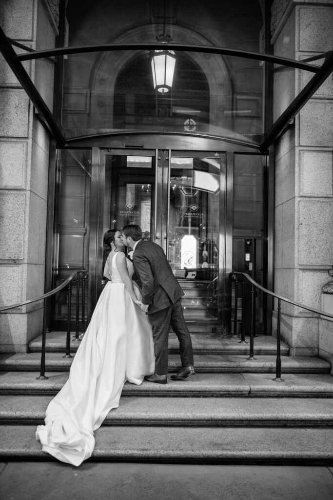 Black and white portrait of bride and groom kissing as they walk up the steps at the University Club of New York photographed by New York City wedding photographer Kat Harris.